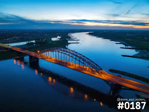 Luchtfoto van de verlichte Waalbrug in Nijmegen na zonsondergang, met oranje lampen die weerkaatsen in de blauwe Waal