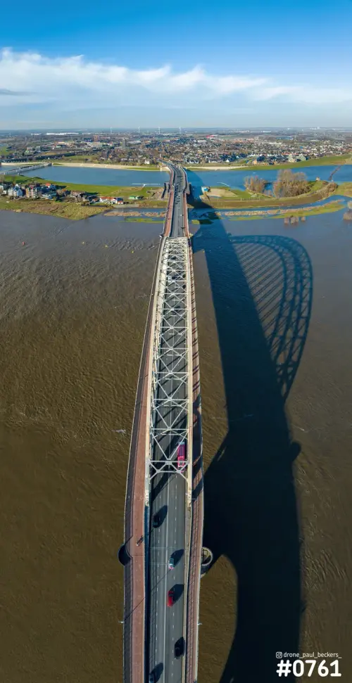 Verticale luchtfoto van de Waalbrug in Nijmegen met de lange schaduw van de boog op het bruine water van de Waal