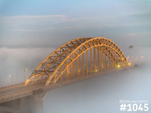 Luchtfoto van de Waalbrug in Nijmegen, de stalen boog goudkleurig verlicht en het brugdek half verdwenen in dikke mist