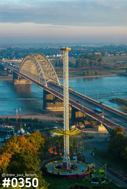 Luchtfoto van de Waalbrug in Nijmegen met een hoge kermisattractie op de voorgrond en herfstbomen langs de Waal