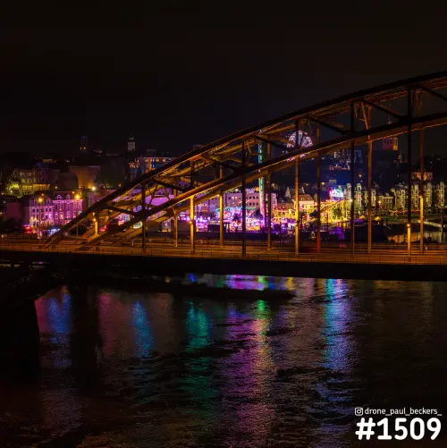 Luchtfoto van de Waalbrug in Nijmegen bij nacht, met kermisverlichting op de Waalkade die weerspiegelt in de Waal