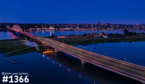 Luchtfoto van de Waalbrug bij Lent in blauw avondlicht, met de verlichte Waalkade en skyline van Nijmegen aan de overkant