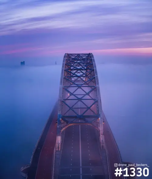 Luchtfoto van de Waalbrug in Nijmegen in schemerlicht, met mist rondom de stalen boog en een lege rijbaan