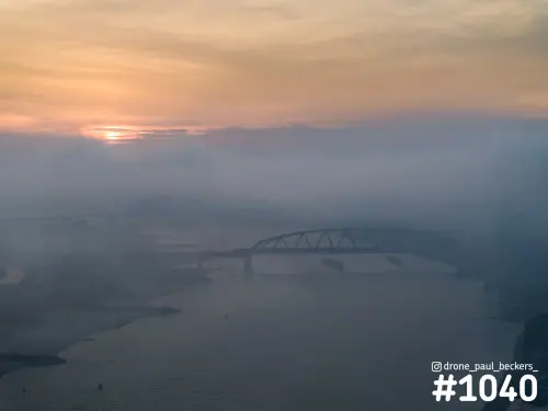Luchtfoto van de Waalbrug in Nijmegen in dichte mist, met laagstaande oranje zon en twee schepen op de Waal