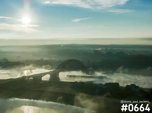 Luchtfoto van de Waalbrug in Nijmegen bij laagstaande zon, met mist boven de Waal en een vrachtschip op het water