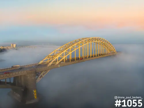 Luchtfoto van de Waalbrug in Nijmegen met gele boog in warm ochtendlicht, terwijl mist de rivier de Waal bedekt