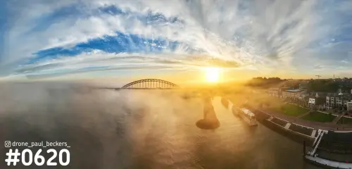 Panorama van de Waalbrug in Nijmegen met laagstaande zon, mist boven de Waal en huizen met een schip aan de Waalkade