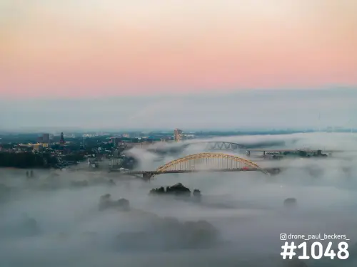 Luchtfoto van de Waalbrug in Nijmegen met laaghangende mist over de Waal en een roze ochtendhemel erboven