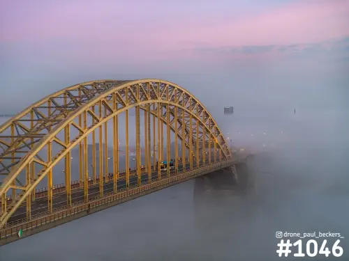 Luchtfoto van de gele boogbrug Waalbrug in Nijmegen in dichte mist, met roze en paarse lucht boven de rivier de Waal