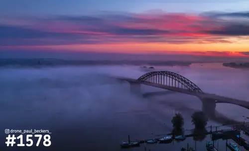Luchtfoto van de Waalbrug in Nijmegen in de schemering, met laaghangende mist boven de Waal en een rood-paarse lucht