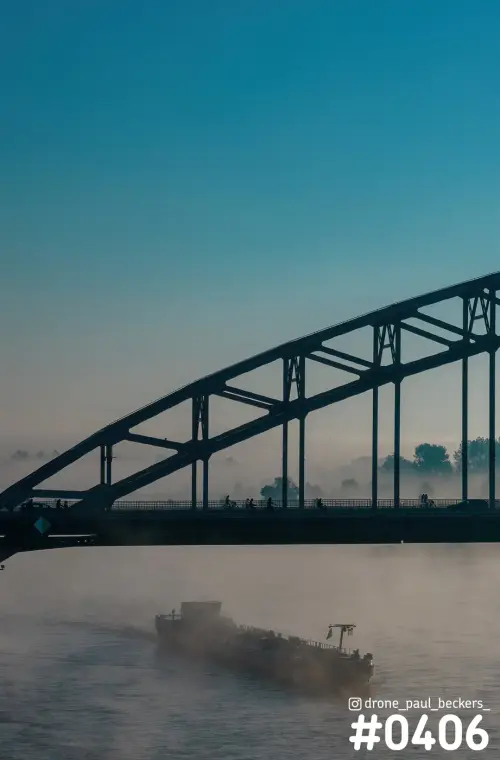 Waalbrug in Nijmegen als silhouet tegen een blauwe lucht, met een binnenvaartschip in de mist op de rivier de Waal