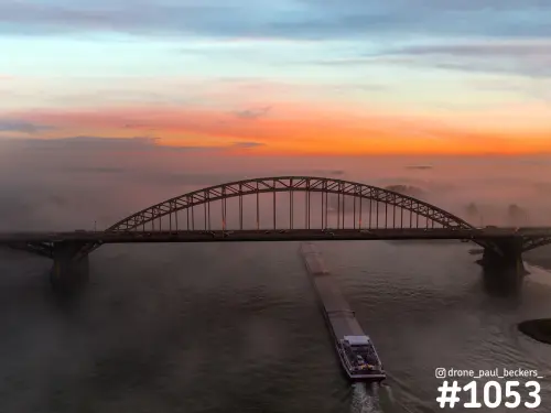 Luchtfoto van de Waalbrug in Nijmegen met mist boven de Waal en een vrachtschip op de rivier, onder een oranje gloed
