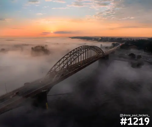 Luchtfoto van de Waalbrug in Nijmegen bij zonsopkomst, met mist over de Waal en verkeer op de boogbrug
