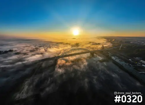 Luchtfoto van de Waalbrug in Nijmegen bij zonsopkomst, met laaghangende mist boven de rivier de Waal en een gouden horizon