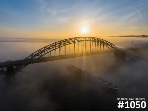 Luchtfoto van de Waalbrug in Nijmegen bij zonsopkomst, met laaghangende mist en een binnenvaartschip op de Waal
