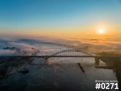 Luchtfoto van de Waalbrug in Nijmegen bij zonsopkomst, met een binnenvaartschip op de Waal en mist boven de Ooijpolder
