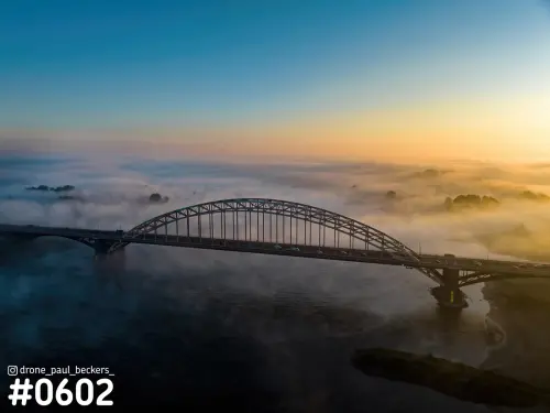 Luchtfoto van de Waalbrug in Nijmegen bij zonsopkomst, met mist boven de Waal en boomtoppen die erdoorheen steken