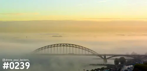 Luchtfoto van de Waalbrug in Nijmegen in dikke mist, met een gouden zonsopkomst boven de rivier de Waal