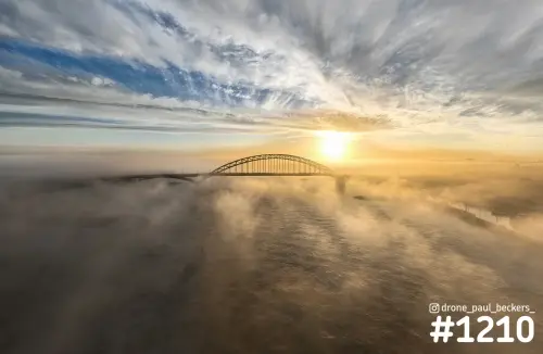 Luchtfoto van de Waalbrug in Nijmegen bij zonsopkomst, met laaghangende mist boven de Waal en de zon naast de boog