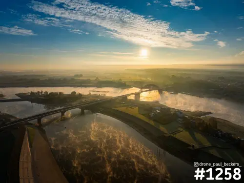 Luchtfoto van de Waalbrug in Nijmegen bij zonsopkomst, met mistslierten boven de Waal en de zon laag boven de horizon