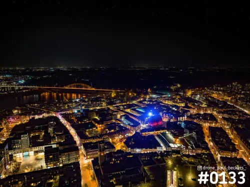 Luchtfoto van Nijmegen bij nacht met de verlichte Waalbrug, de Waal en een blauw en rood verlicht gebouw in het centrum