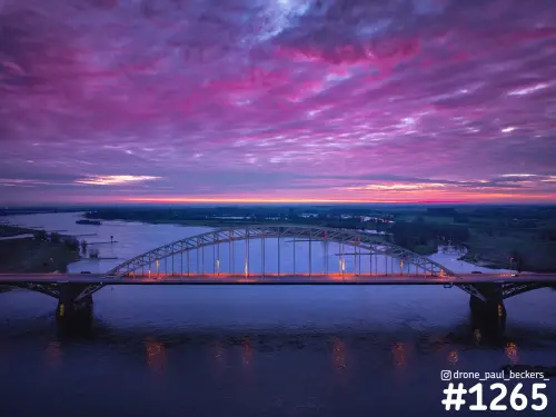 Luchtfoto van de Waalbrug in Nijmegen in de schemering, met roze en paarse wolken boven de Waal en de uiterwaarden