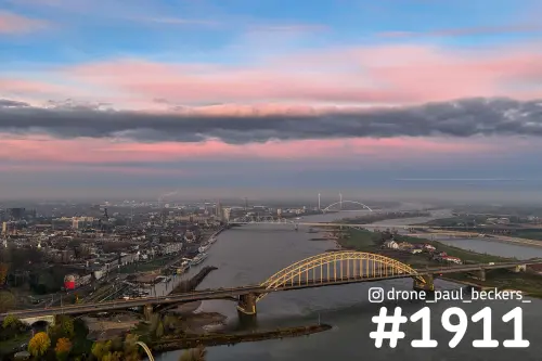 Luchtfoto van de Waalbrug in Nijmegen bij schemering, met roze wolken boven de Waal en De Oversteek in de verte