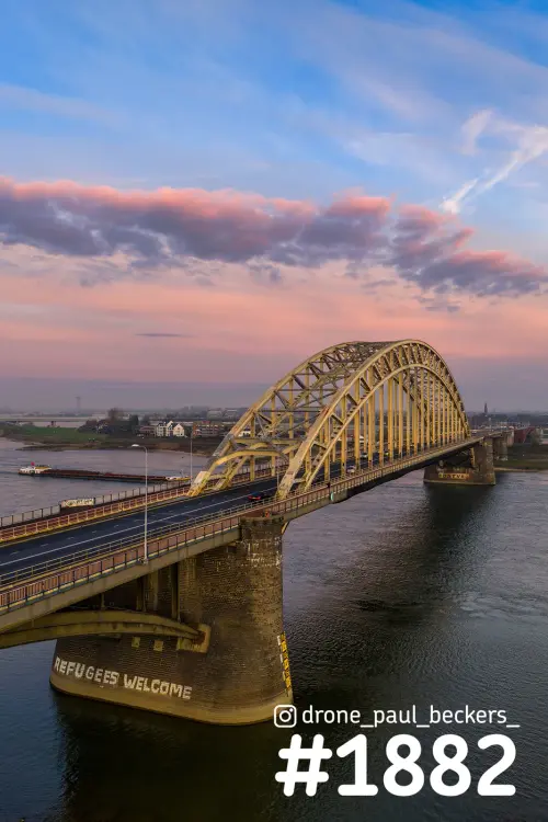 Luchtfoto van de Waalbrug in Nijmegen met roze wolken en warm licht op de gele boogconstructie boven de Waal