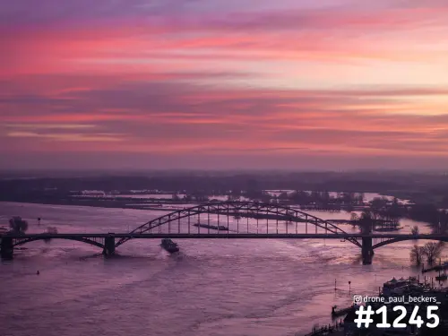 Luchtfoto van de Waalbrug in Nijmegen in roze ochtendlicht, met een binnenvaartschip op de Waal en hoog water