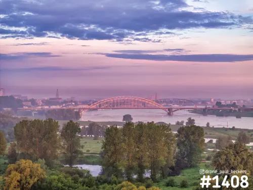 Luchtfoto van de Waalbrug in Nijmegen bij zonsopkomst, met roze lucht boven de Waal en bomen in de Ooijpolder