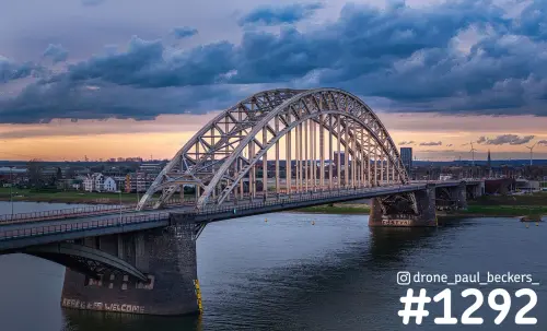 Luchtfoto van de Waalbrug in Nijmegen in de schemering, met donkere wolken en een oranje gloed boven de Waal