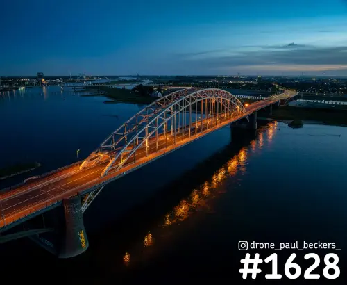Luchtfoto van de stalen bogen en stenen brugpijler van de Waalbrug in Nijmegen, met schemerlucht en De Oversteek in de verte