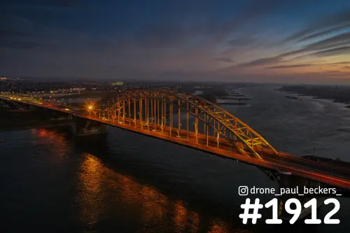 Luchtfoto van de verlichte boogbrug Waalbrug in Nijmegen bij schemering, met oranje reflecties op de Waal