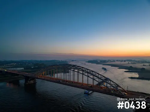 Luchtfoto van de Waalbrug in Nijmegen in de schemering, met oranje gloed aan de horizon en een schip op de Waal