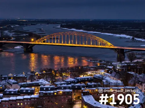 Luchtfoto van de verlichte Waalbrug in Nijmegen in de avondschemering, met sneeuw op de daken en langs de Waal