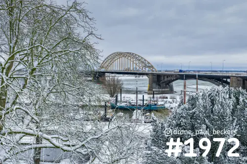 Waalbrug in Nijmegen onder een grijze winterlucht, gezien door besneeuwde takken met een boot in de Lindenberghaven