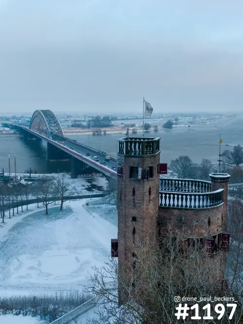 Luchtfoto van de Waalbrug in Nijmegen in de sneeuw, met op de voorgrond een bakstenen toren met vlag aan de Waal