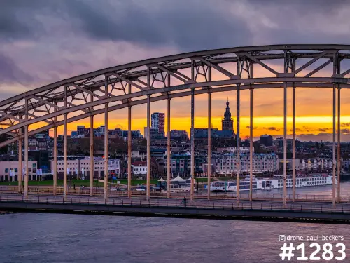 Boog van de Waalbrug in Nijmegen met Stevenskerk en Waalkade bij zonsondergang, oranje lucht en een rivierschip