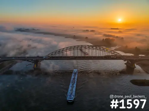 Luchtfoto van de Waalbrug in Nijmegen bij zonsopkomst, met een tanker op de Waal en mist boven de Ooijpolder