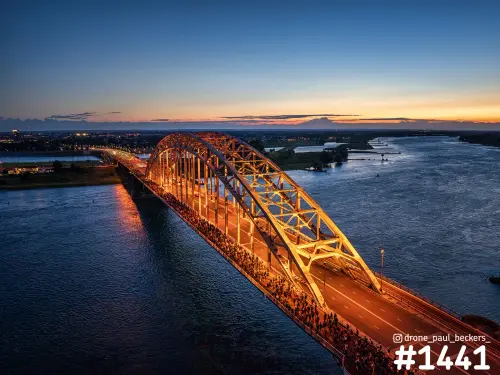 Luchtfoto van de verlichte Waalbrug in Nijmegen bij schemering, met een lange rij mensen op het wegdek en oranje gloed boven de Waal