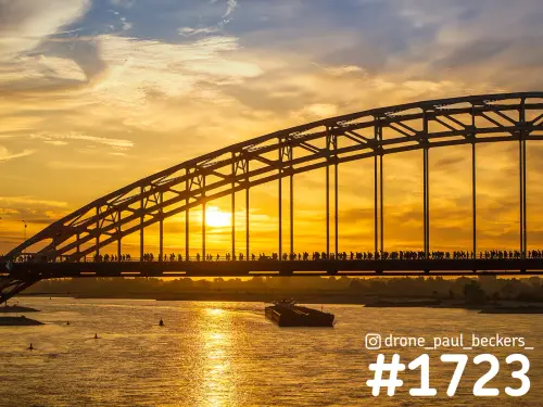 Waalbrug in Nijmegen met silhouetten van Vierdaagse-lopers tegen een gouden lucht, met een binnenvaartschip op de Waal