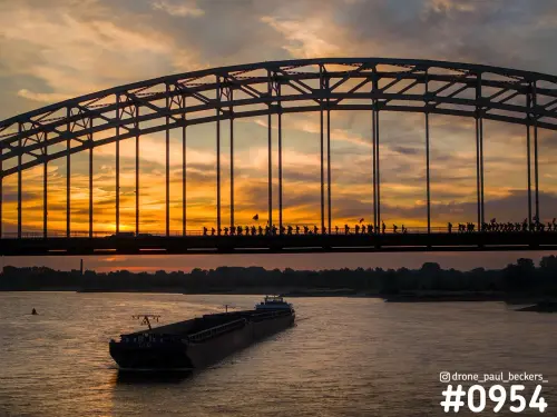 Wandelaars van de Vierdaagse als silhouet op de Waalbrug in Nijmegen, met oranje gloed en een binnenvaartschip op de Waal
