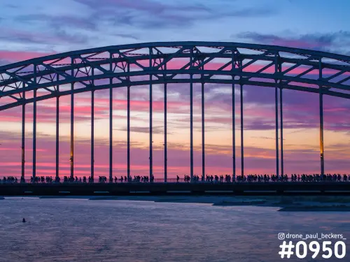 Waalbrug in Nijmegen met silhouetten van Vierdaagse-lopers voor een roze en paarse schemerlucht, over de Waal