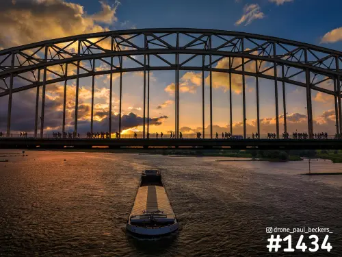 Luchtfoto Waalbrug Nijmegen bij zonsondergang, binnenvaartschip op de Waal en wandelaars als silhouetten op de brug