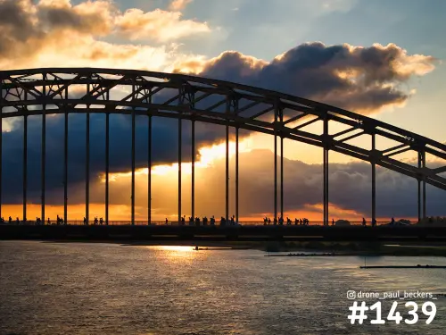 Waalbrug in Nijmegen als silhouet bij zonsondergang, met wandelaars van de Vierdaagse op het brugdek en de rivier de Waal eronder