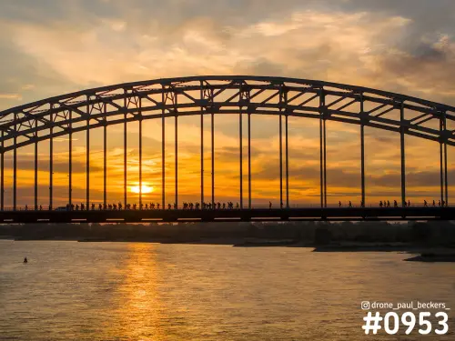 Wandelaars van de Vierdaagse als silhouetten op de Waalbrug in Nijmegen bij zonsopkomst, met oranje lucht boven de Waal