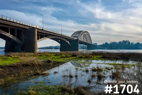 Waalbrug in Nijmegen vanaf de oever, met de boogbrug weerspiegeld in een plas tussen riet en gras onder wolkenlucht