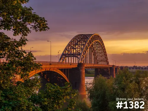 Waalbrug in Nijmegen met oranje en paarse avondlucht, gezien tussen het groene bladerdek van bomen aan de Waal