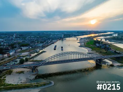 Luchtfoto van de Waalbrug in Nijmegen bij zonsondergang, met schepen op de Waal en De Oversteek in de verte