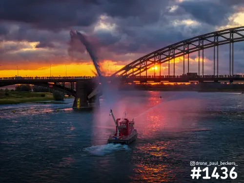 Luchtfoto van de Waalbrug in Nijmegen bij zonsondergang, met een rode boot die water spuit en mensen op de brug